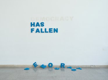 Wand mit aufgehängten Buchstaben die die Wörter "Has Fallen" bilden. Auf dem Boden liegen die Buchstaben für das Wort "Democracy"