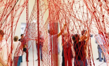 In einem hohen Raum stehen Menschen unter einem roten Netz, das an der Decke hängt. Das Netz stellt einen Baumstamm dar mit riesigen Wurzeln und an diesen Wurzeln knüpfen die Personen weiter.