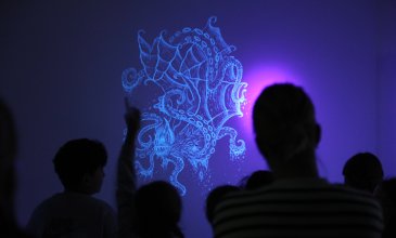 Schatten von Menschen vor einem UV-Licht leuchtenden Wandbild eines Fantasiewesens