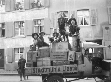 Auf der Schwarzweissfotografie von 1927 ist ein alter Lastwagen mit der Aufschrift "Stadtmühle Liestal" zu sehen, auf dessen Ladefläche sich Obstkisten stapeln. Auf und um den Wagen stehen und sitzen acht Kinder und Jugendliche in Pfadiuniform, die meisten grinsen stolz in die Kamera. Im Hintergrund sieht man Häuser, eine Frau schaut aus dem Fenster.