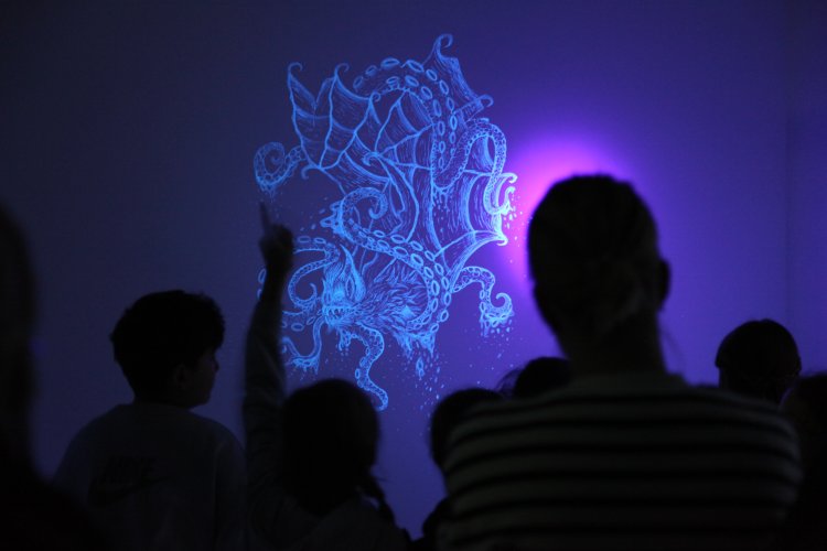 Ein von UV-Licht beleuchtetes Wandbild in Form eines Monsters, davor sind Schatten von Menschen