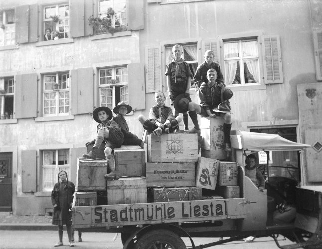 Auf der Schwarzweissfotografie von 1927 ist ein alter Lastwagen mit der Aufschrift "Stadtmühle Liestal" zu sehen, auf dessen Ladefläche sich Obstkisten stapeln. Auf und um den Wagen stehen und sitzen acht Kinder und Jugendliche in Pfadiuniform, die meisten grinsen stolz in die Kamera. Im Hintergrund sieht man Häuser, eine Frau schaut aus dem Fenster.