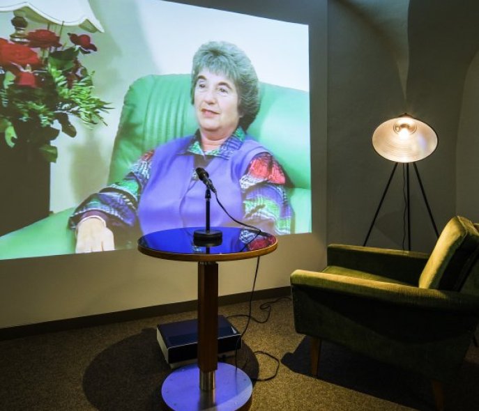 On a screen, you can see an older woman speaking; in the foreground is living room furniture.
