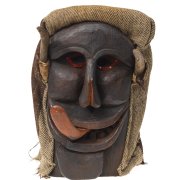 Maske aus dunkelbraunem Holz mit rötlichen Augen, gespaltener Nase, grossem leicht geöffnetem Mund, aus dem die Zunge links herausgestreckt ist. Anstatt Haare trägt die Maske einen Sack.