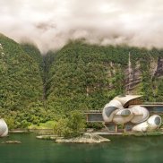 Das Bild zeigt eine idyllische Fjordlandschaft mit steilen, dicht bewaldeten Berghängen, deren Spitzen in dichte Wolken gehüllt sind. Im Vordergrund liegt ein ruhiger See, auf dem mehrere futuristisch-organisch geformte Gebäude schwimmen oder am Ufer stehen. Die Architektur erinnert an große, geschwungene Kapseln mit glatten, weißen Oberflächen und großen Panoramafenstern. Die Szene wirkt wie eine Mischung aus Naturidylle und moderner Sci-Fi-Architektur.