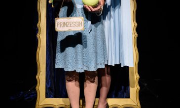 Zwei Frauen, eine mit Krönchen auf dem Kopf, die andere mit einem Schild, auf dem Prinzessin steht, stehen in einem goldenen Rahmen. Eine Handpuppe in Gestalt eines Frosches blickt neugierig auf die goldene Kugel in der Hand  einer der Prinzessinnen.