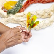 Zwei Hände knüpfen gelb-rot-grüne Federn zusammen.