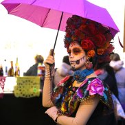 Catrina, üppig geschminkte Frau mit Rosen in den Haaren und sehr farbigem Kleid in rosa, orange, elb, blau, grün