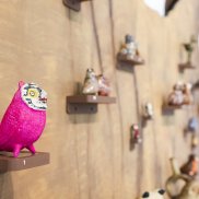 An einem Baumstamm an einer Wand stehen Eulen auf kleinen Podest. Im Vordergrund ist eine pinke Eule zu sehen.