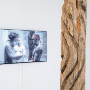Dhulu, der Baumstamm, steht vor einem Bildschirm auf welchem eine indigene Persönlichkeit ein Ritual mit einer Kuratorin des Museums abhält