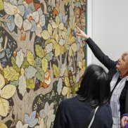 Anna Schmid und der Wandteppich mit dem Muttenzer Wald Anna Schmid zeigt in der Weihnachtsausstellung 2024 auf einen Wandteppich des Muttenzer Waldes,.Neben ihr stehen zwei Besucher*innen