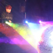 Djane an den Turntables und vor ihr strahlen die Lichter in Regenbogenfarben auf eine Discokugel und alles reflektiert farbenfroh