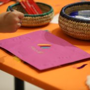 Auf einem orangen Tisch liegt ein pinkes Blatt, auf dem eine Kinderhand zeichnet.