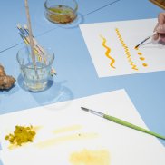 Auf blauem Tisch liegen weisse Blätter, auf denen mit gelber Farbe gemalt wird.