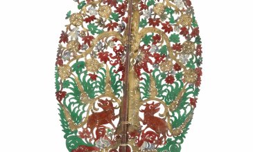 Ovale gestanzte Form mit Stiel, auf der ein Baum erkennbar ist mit grünen Blättern und roten Blüten und zwei rote Tiere am unteren Ende des Stammes.