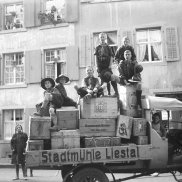 Auf der Schwarzweissfotografie von 1927 ist ein alter Lastwagen mit der Aufschrift "Stadtmühle Liestal" zu sehen, auf dessen Ladefläche sich Obstkisten stapeln. Auf und um den Wagen stehen und sitzen acht Kinder und Jugendliche in Pfadiuniform, die meisten grinsen stolz in die Kamera. Im Hintergrund sieht man Häuser, eine Frau schaut aus dem Fenster.