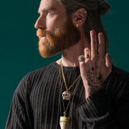 Ein Mann mit langen Haaren und Bart trägt einen schwarzen Pulli, Ohrringe, mehrere Ketten, Ringe und ein Tattoo am Handrücken.