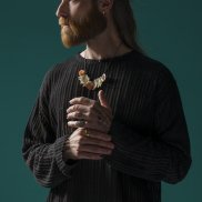 Ein Mann mit langen Haaren und Bart trägt einen schwarzen Pulli, Ohrringe, mehrere Ringe und eine Kette aus Zähnen und Muscheln.