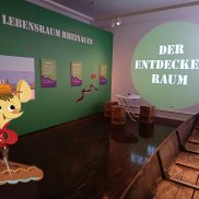 Blick in die Ausstellung, © Maria Karipidou/Stadtmuseum zu sehen sind Infotafeln im Museumsraum mit Sitzgelegenheit, dem Nachen und im Vordergrund die Maus einkopiert