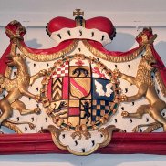 Wappen mit Hermelinmantel und Krone umrahmt, das Wappen selbst zeigt verschiedene Bilder wie Karo oder Streifen, Tiere und Blumen