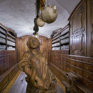 Holzgeschnitzte Figur auf einem Apothekentisch, links und rechts das Mobiliar der Innsbrucker Hofapotheke. An der Decke hängt ein ausgestopfter Kugelfisch