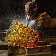 un vase en cristal ambre avec des motifs de boules est en train d'être taillé par une main qui tient un outil et une autre qui tient un jet d'eau