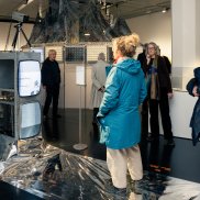 Besucher:innen der Ausstellung stehen vor zwei übereinander gestapelten Fernsehern, auf denen ein Video läuft.