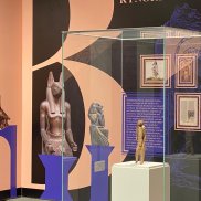 Farbfotografie: Ansicht eines Ausstellungsraumes mit farbiger Wantapete. Im Zentrum eine Vitrine, in der auf einem weißen Sockel eine braune Figur steht.