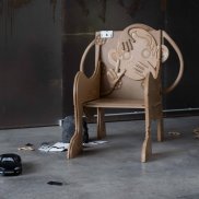 Simone Eisele, „Good Morning, Mickey!“, Installationsansicht Motorworld Manufaktur Metzingen (Detail), 2020. Aluminium, Buche, MDF, Pappe, Edding, Stoff, Füllwatte, Lack, Brotpapier, Perspex, Rock Speaker. Foto: Simone Eisele. © the artist. Installation eines geschnitzen Stuhls, Lampe, Spielzeugauto, Papier, und Trümmer