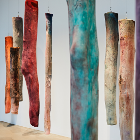 Zu sehen ist ein Ausschnitt einer Rauminstallation. Bunte, holzartige Körper hängen von der Decke, die an Baumstämme erinnern können.