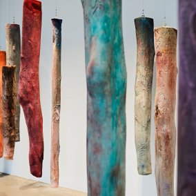 Das Bilde zeigt einen Blick in die Installation des Werkes Mothers leg's der Künstlerin Kaari Upson. An Drahtseil hängen baumstammartige Elemente von der Decke. Bei näherem betrachten, erinnern sie an ein Ausschnitt von Beinen mit Knien. Verfremdet wird die Darstellung durch das bunte Erscheinungsbild der Elemente.