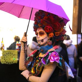 Catrina, üppig geschminkte Frau mit Rosen in den Haaren und sehr farbigem Kleid in rosa, orange, elb, blau, grün