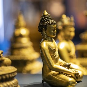 Das Foto zeigt einen sitzenden Buddha mit heller Hautfarbe, der goldene Kleidung trägt. Er sitzt auf einem roten Sockel.