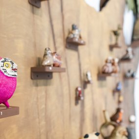 Auf einem Baumstamm an einer Wand stehen viele kleine Eulen auf kleinen Podesten. Im Vordergrund sitzt eine pinke Eule.