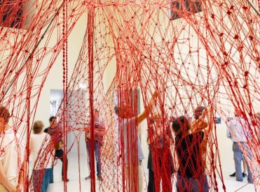 In einem hohen Raum stehen Menschen unter einem roten Netz, das an der Decke hängt. Das Netz stellt einen Baumstamm dar mit riesigen Wurzeln und an diesen Wurzeln knüpfen die Personen weiter.