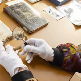 Eine Person mit weißen Handschuhen untersucht mit einer Lupe historische Objekte und Fotos auf einem Tisch. Die Szene wirkt wie Archiv- oder Museumsarbeit.