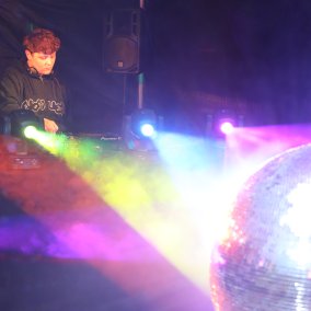 Djane an den Turntables und vor ihr strahlen die Lichter in Regenbogenfarben auf eine Discokugel und alles reflektiert farbenfroh