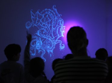 Schatten von Menschen vor einem UV-Licht leuchtenden Wandbild eines Fantasiewesens