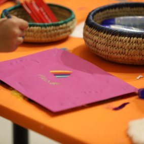 Auf einem orangen Tisch liegt ein pinkes Blatt, auf dem eine Kinderhand zeichnet.