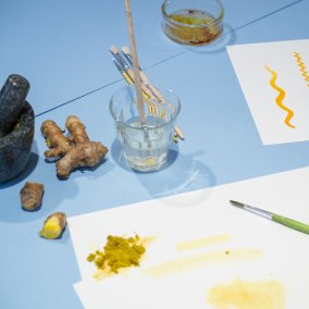 Auf blauem Tisch liegen weisse Blätter, auf denen mit gelber Farbe gemalt wird.