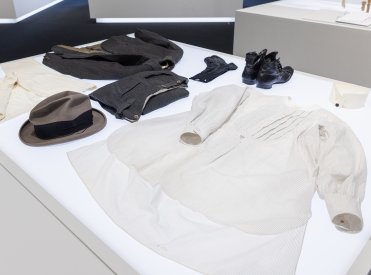 Auf einem weissen Ausstellungstisch liegt eine Herrenausstattung mit Jacke, Hosen, Hemd, Kragen, Weste, Unterhosen, Socken, Filzhut, Schuhen aus Basel, Schweiz  von 1924