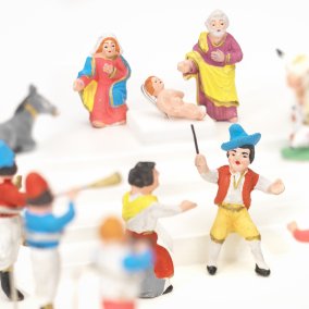 Farbige Krippenfiguren, im Hintergrund Maria, Jesus und Josef, daneben eine weisse Kuh und ein grauer Esel, davor eine Gruppe von Musizierenden.