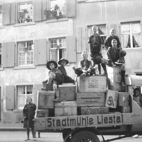 Auf der Schwarzweissfotografie von 1927 ist ein alter Lastwagen mit der Aufschrift "Stadtmühle Liestal" zu sehen, auf dessen Ladefläche sich Obstkisten stapeln. Auf und um den Wagen stehen und sitzen acht Kinder und Jugendliche in Pfadiuniform, die meisten grinsen stolz in die Kamera. Im Hintergrund sieht man Häuser, eine Frau schaut aus dem Fenster.