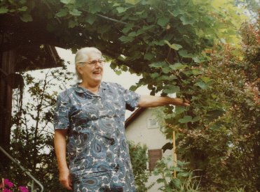 Zu sehen ist eine ältere, lachende Frau mit weissem Haar und Brille umgeben von grünen Pflanzen.