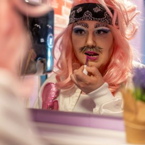 Eine geschminkte Person mit pinker Perücke sieht in einen Spiegel und trägt Lippenstift auf.