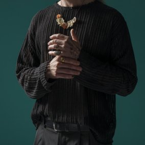 Ein Mann mit langen Haaren und Bart trägt einen schwarzen Pulli, Ohrringe, mehrere Ringe und eine Kette aus Zähnen und Muscheln.