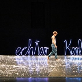 Ein Mann geht barfuß über eine Bühne, hinter ihm eine Leuchtschrift „ein echter Kerl“