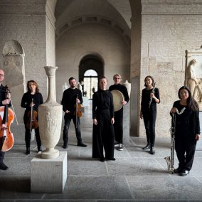 Sieben Musiker*innen stehen mit ihren Instrumenten und schwarz gekleidet in einem historisch anmutenden Raum.