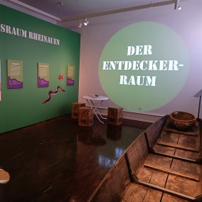 zu sehen sind Infotafeln im Museumsraum mit Sitzgelegenheit, dem Nachen und im Vordergrund die Maus einkopiert