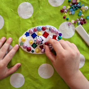 Kinderhände verzieren eine Tonscheibe mit bunten Glasperlen